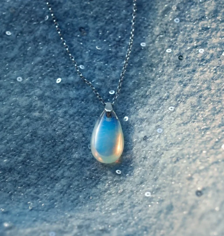 A teardrop-shaped opalescent pendant on a silver chain rests on a soft, light blue fabric adorned with small, sparkling sequins.