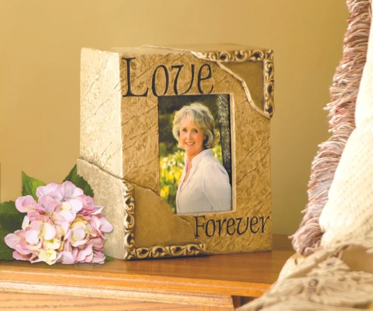 A decorative urn with a photo of an older woman smiling, framed by the words Love at the top and Forever at the bottom. A pink hydrangea and a fringed blanket are nearby on the table.